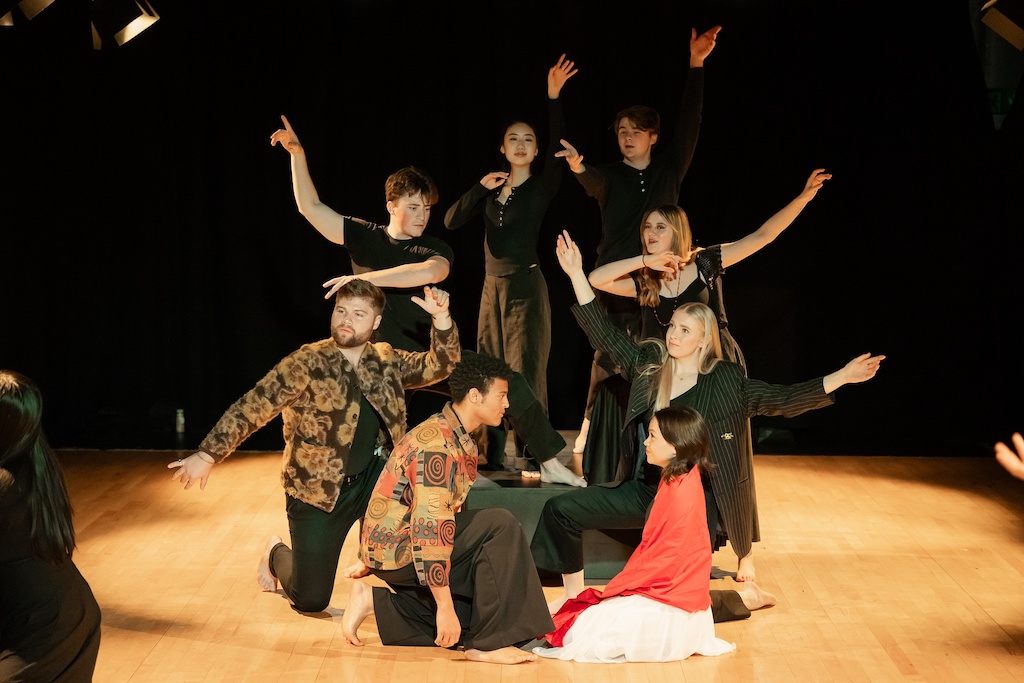 Two actors kneel on stage facing each other while another group of actors with arms outstretched stands or kneels behind them.